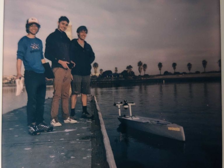 Three people stand on a pier next to an autonomous boat